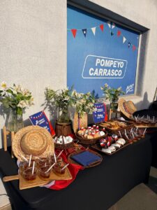 Mesa con empanadas, dulces y mote con huesillos, decorada con banderas chilenas y sombreros, frente al letrero de Pompeyo Carrasco.